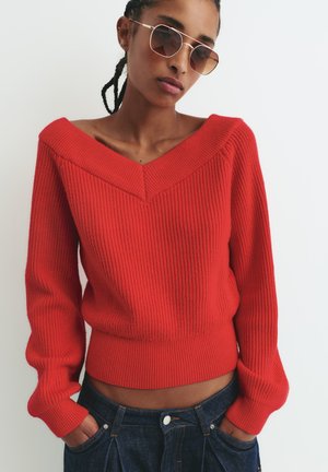 Maglione a coste rosso con scollo a V, dotato di maniche lunghe e vita aderente. Abbinato a jeans scuri, che mostrano uno stile casual.