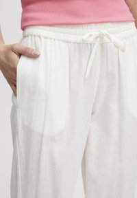 Pantalon en lin blanc avec une taille à cordon, des poches latérales et une coupe décontractée. Le tissu a une texture lisse et une apparence légère et aérienne.