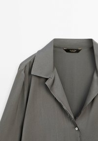 Chemise grise à col avec une coupe décontractée, présentant des bords effilochés près de la zone des boutons et une étiquette de marque à l'intérieur du col.