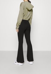 Woman wearing black flared pants, light olive cropped hoodie, and white sneakers, standing against a plain white background, back facing viewer.