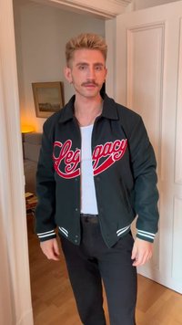 Man with blond hair and mustache wearing a black jacket with "Legacy" in red, standing indoors near a doorway with a painting in the background.