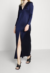 Navy satin long dress with long sleeves, V-neckline, and thigh-high side slit. Paired with black open-toe block-heeled sandals.