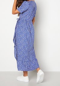 Modré květinové šaty s krátkými rukávy, volánkovým lemem a bílým květinovým vzorem; spárované s bílými teniskami. Hladká textilie.