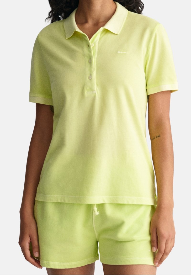 Femme portant un polo à manches courtes de couleur jaune clair et un short assorti, debout les bras détendus contre un fond uni.