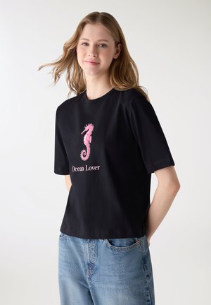 Giovane donna con capelli biondi che indossa una maglietta nera con un cavalluccio marino rosa e la scritta "Ocean Lover", abbinata a jeans blu.