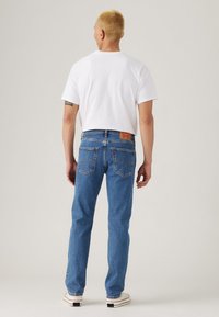 Personne aux cheveux blonds courts portant un t-shirt blanc, un jean bleu et des baskets blanches, debout de dos sur un fond uni.