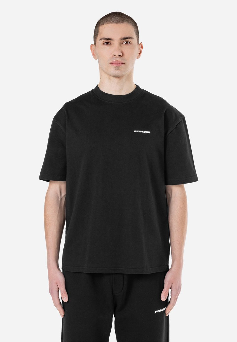 Junger Mann mit kurzen Haaren, trägt ein schwarzes Pegador T-Shirt und passende schwarze Hose, steht vor weißem Hintergrund.