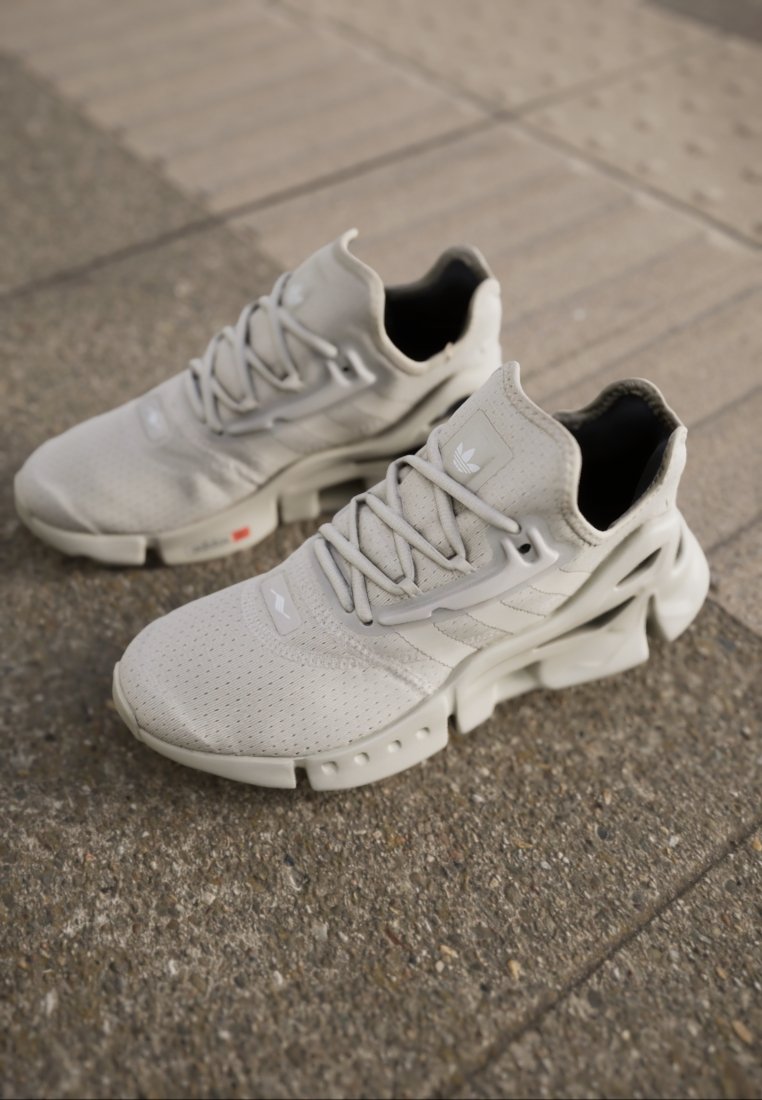 Par de zapatillas deportivas Adidas de color gris claro con tela perforada, diseño intrincado en la suela y cordones descansando sobre un pavimento texturizado.