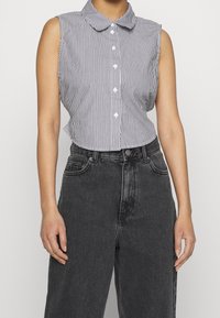 Ärmlös, kragig skjorta i svart och vitt med vertikala ränder, med knapprad framtill och en kortad nederkant, bärs med mörka högmidjade jeans.