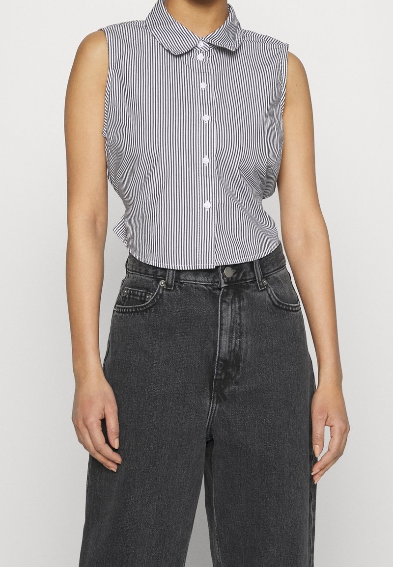 Ärmlös, kragig skjorta i svart och vitt med vertikala ränder, med knapprad framtill och en kortad nederkant, bärs med mörka högmidjade jeans.