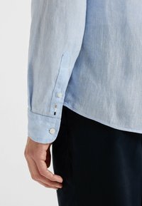 Jasnoniebieska lniana koszula z guzikowym mankietem, charakteryzująca się subtelną fakturą i kontrastującymi akcentami nici w pobliżu guzików. Widoczne czarne spodnie.