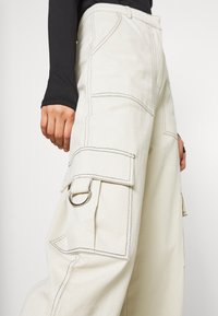 Beige cargo kalhoty s kontrastním černým šitím, velkými bočními kapsami a detaily ze stříbrného kovu. Volný střih.