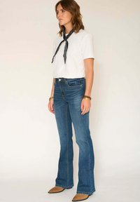 Femme debout portant un t-shirt blanc, un jean évasé bleu, des chaussures marron pointues et une écharpe noire nouée autour du cou.