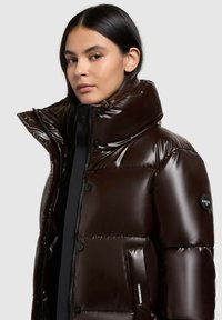Brown puffer jacket with glossy finish, high collar, button closures, and side pocket. Accented with a logo badge on the sleeve.