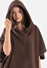 Brązowy polarowy ponczo z dużym kapturem i szerokimi rękawami. Posiada kieszeń z przodu oraz ribbowy wzór. Casualowy, luźny krój.