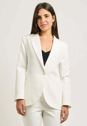 Donna con lunghi capelli scuri che indossa una giacca bianca sopra una maglietta nera, in piedi davanti a uno sfondo chiaro e uniforme.