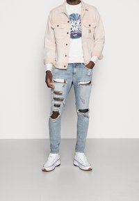 Chaqueta de mezclilla rosa claro sobre una camiseta gráfica blanca, combinada con jeans claros rasgados y zapatillas blancas. Desgastes notables en los jeans.