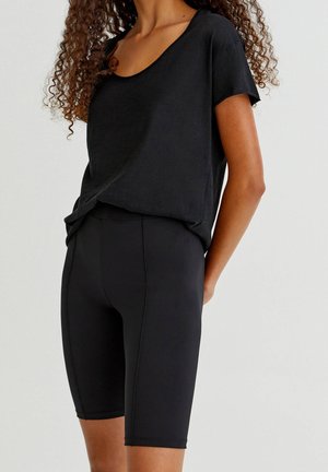 Vrouw draagt een zwart T-shirt met korte mouwen en ronde hals en zwarte aansluitende knielange fietsshorts, staand tegen een effen achtergrond.