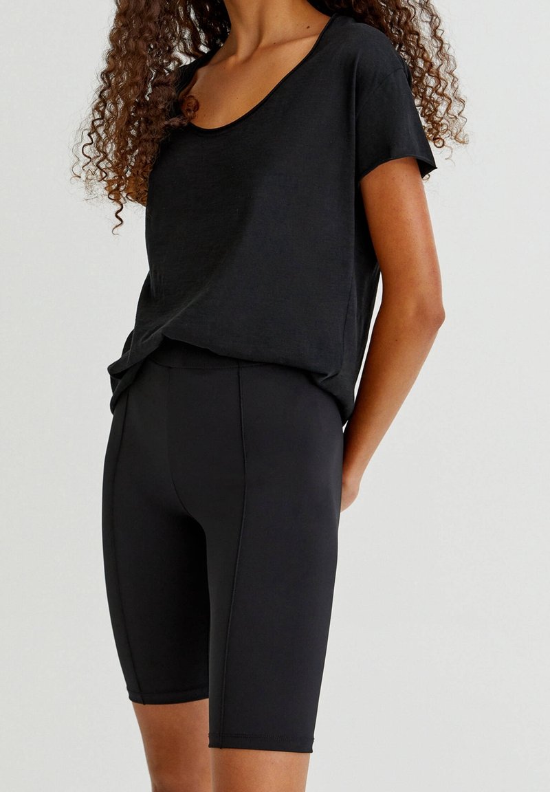Femme portant un t-shirt noir à manches courtes et col rond ainsi que des shorts de cyclisme noirs ajustés jusqu’aux genoux, debout devant un fond uni.