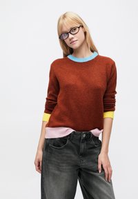 Maglione marrone lavorato a maglia con colletto azzurro chiaro, polsini gialli e orlo rosa, abbinato a jeans in denim grigio a vita alta.