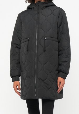 Veste matelassée noire à capuche avec fermeture éclair frontale, poches zippées avec cordons, et poignets élastiques, portée par une personne debout.