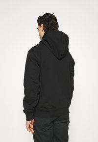 Muž s kudrnatými vlasy, oblečený v jednoduché černé mikině s kapucí a černých kalhotách, stojící zády k bílé stěně.