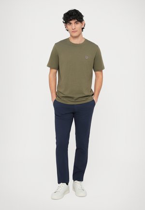 Olivovo zelené bavlnené tričko s krátkymi rukávmi, tmavomodré nohavice a biele tenisky. Malý vyšívaný detail na hrudi trička.