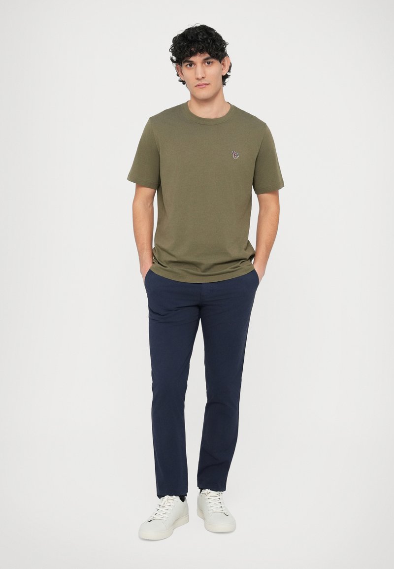 Tricou de bumbac verde măsliniu cu mâneci scurte, pantaloni bleumarin și adidași albi. Detaliu mic brodat pe pieptul tricoului.