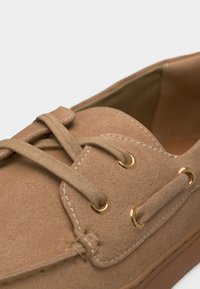 Sapatilha náutica em camurça castanha clara com atacadores bege passados por ilhóses dourados e detalhes de costura branca visível.