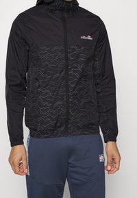 Svart vindjacka med dragkedja fram, elastiska manschetter och ett vågmönster i grått över bröstområdet. Har en huva.