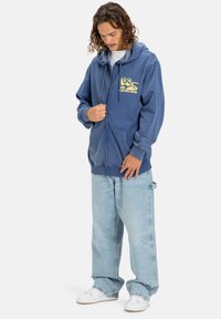Námornícky modrá mikina s kapucňou na zips a klokaním vreckom, s žltým logom na hrudi. Nosená s svetlomodrými širokými džínsami.