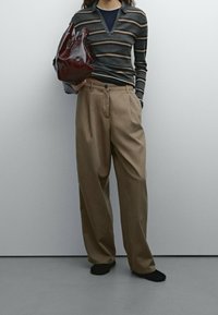 Paski szare i brązowe długie rękawy polo, beżowe spodnie z wysokim stanem, czarne skarpety, trzymający błyszczącą bordową torbę, na neutralnym tle.