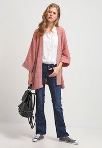 Light pink oversized cardigan made of a textured knit, paired with a white button-up shirt and dark blue flared jeans. Accessories include a black backpack.
