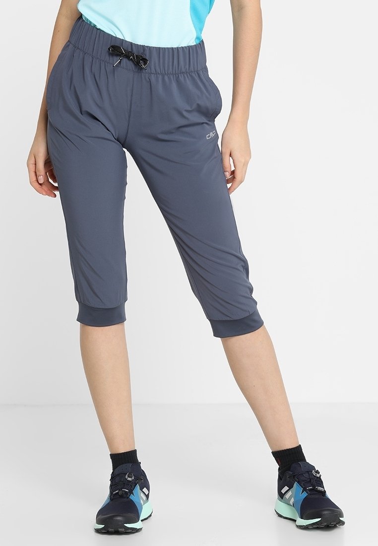 Personne portant un pantalon de sport gris court avec un cordon noir et des baskets foncées avec des accents bleu clair, debout sur un sol blanc.