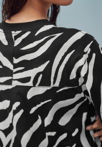 Crna dukserica s izrazitim bijelim uzorkom zebra pruga, s okruglim izrezom i teksturiranim materijalom. Bez bora i prilagođenog kroja.