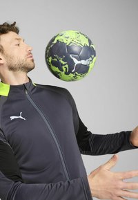 Sort og grå langærmet træningsjakke med lynlås, der har en lys grøn krave. En mønstret fodbold svæver foran bæreren.