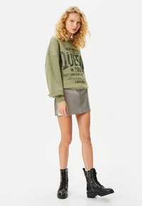 Mujer con cabello rubio rizado, vistiendo un sudadera verde oliva con texto, una falda corta gris y botas negras con cordones, de pie sobre un fondo blanco.