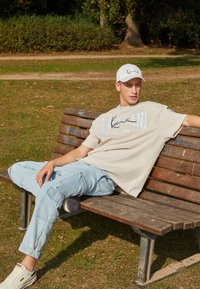T-shirt beige surdimensionné avec texte graphique, pantalon cargo bleu clair et baskets blanches, porté par une silhouette assise sur un banc en bois.