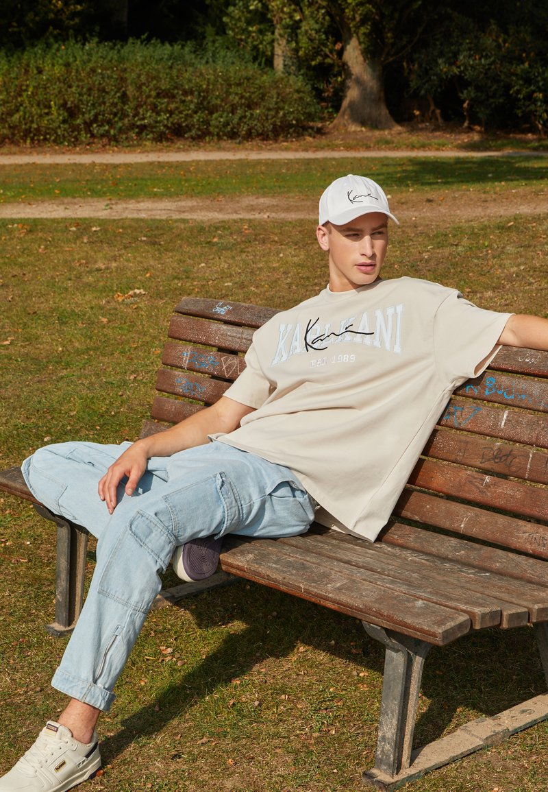 T-shirt beige surdimensionné avec texte graphique, pantalon cargo bleu clair et baskets blanches, porté par une silhouette assise sur un banc en bois.