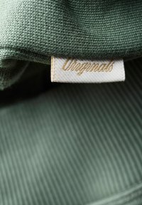 Gros plan d’un tissu tricoté vert avec une étiquette blanche portant le mot « Originals » en texte doré cursif.