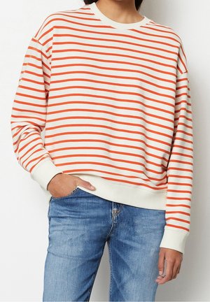 Persoon draagt een wit trui met lange mouwen met horizontale oranje strepen en een blauwe spijkerbroek, met één hand in de zak.