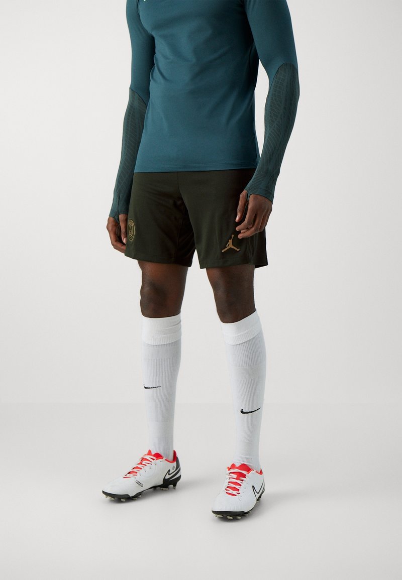 Chemise de sport bleu sarcelle à manches longues, short olive avec des accents dorés, chaussettes blanches et chaussures de football rouge et blanc avec des détails noirs.