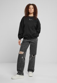 Svart oversized genser med hvitt logo, kombinert med slitte grå vide jeans med rifter og hvite sneakers.