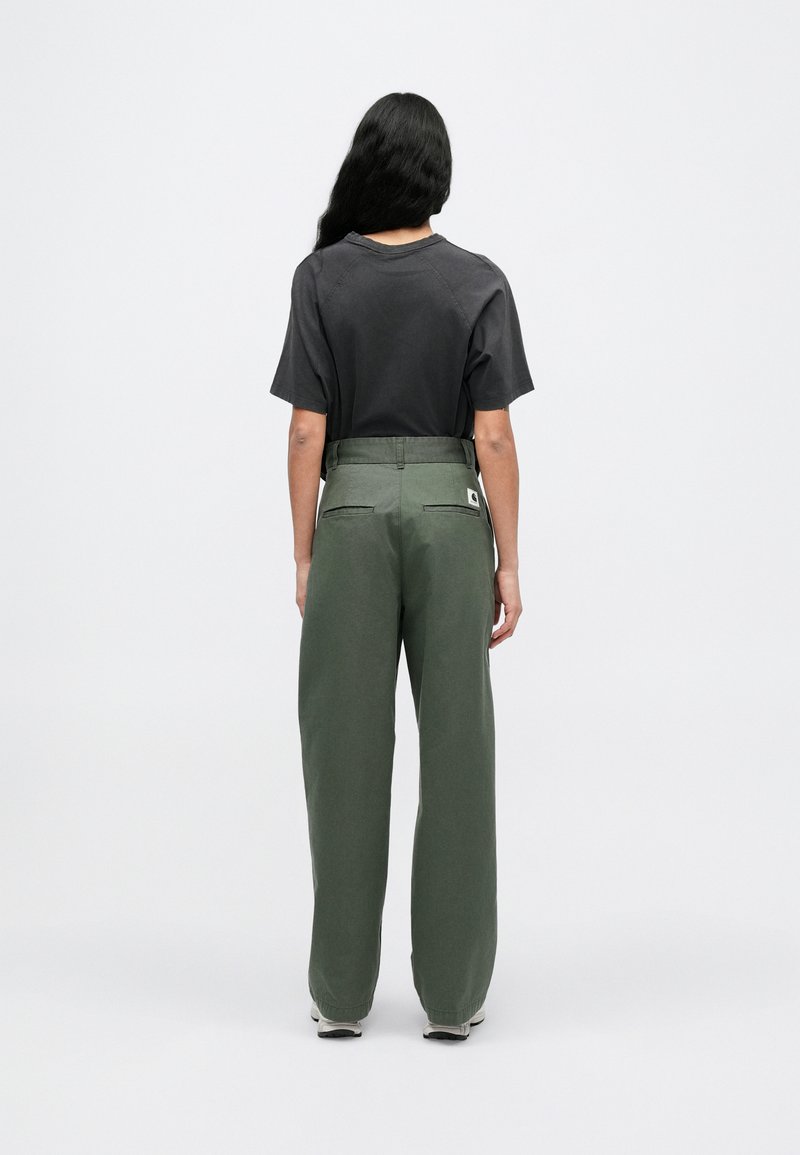 Schwarzes T-Shirt mit kurzen Ärmeln, kombiniert mit grünen, weit geschnittenen Hosen mit Gesäßtaschen und einem Logotag, von hinten vor schlichtem Hintergrund betrachtet.