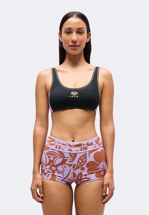 Le modèle est debout, face à l’avant, portant une brassière de sport noire Roxy et un short lilas à motif floral couleur rouille.