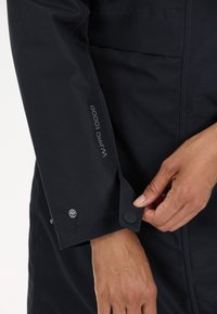 Cappotto nero con etichetta WPRO 10000. Presenta un polsino aderente con chiusura a bottone a pressione e una finitura in tessuto morbido e testurizzato.