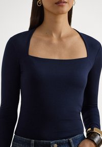 Tengerészkék hosszú ujjú body négyzet alakú kivágással, sima textúrával és testhezálló dizájnnal, arany karkötővel és farmernadrággal párosítva.