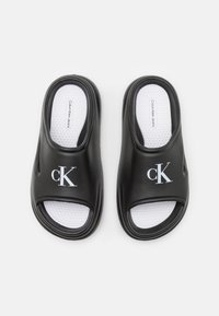 Sandales noires à glissière en plastique lisse avec une semelle intérieure texturée blanche. Présente un logo "cK" blanc sur la bride supérieure.