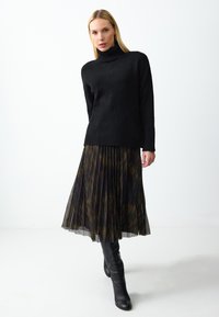Schwarzer gestrickter Rollkragenpullover kombiniert mit einem gefaltenen schwarzen und mehrfarbigen Rock. Model trägt knielange schwarze Lederstiefel.
