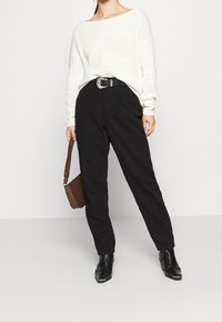 Dikke witte gebreide trui, zwart hooggewaisted broek, zwarte gestructureerde enkellaarzen en een gevlekte bruine schoudertas.